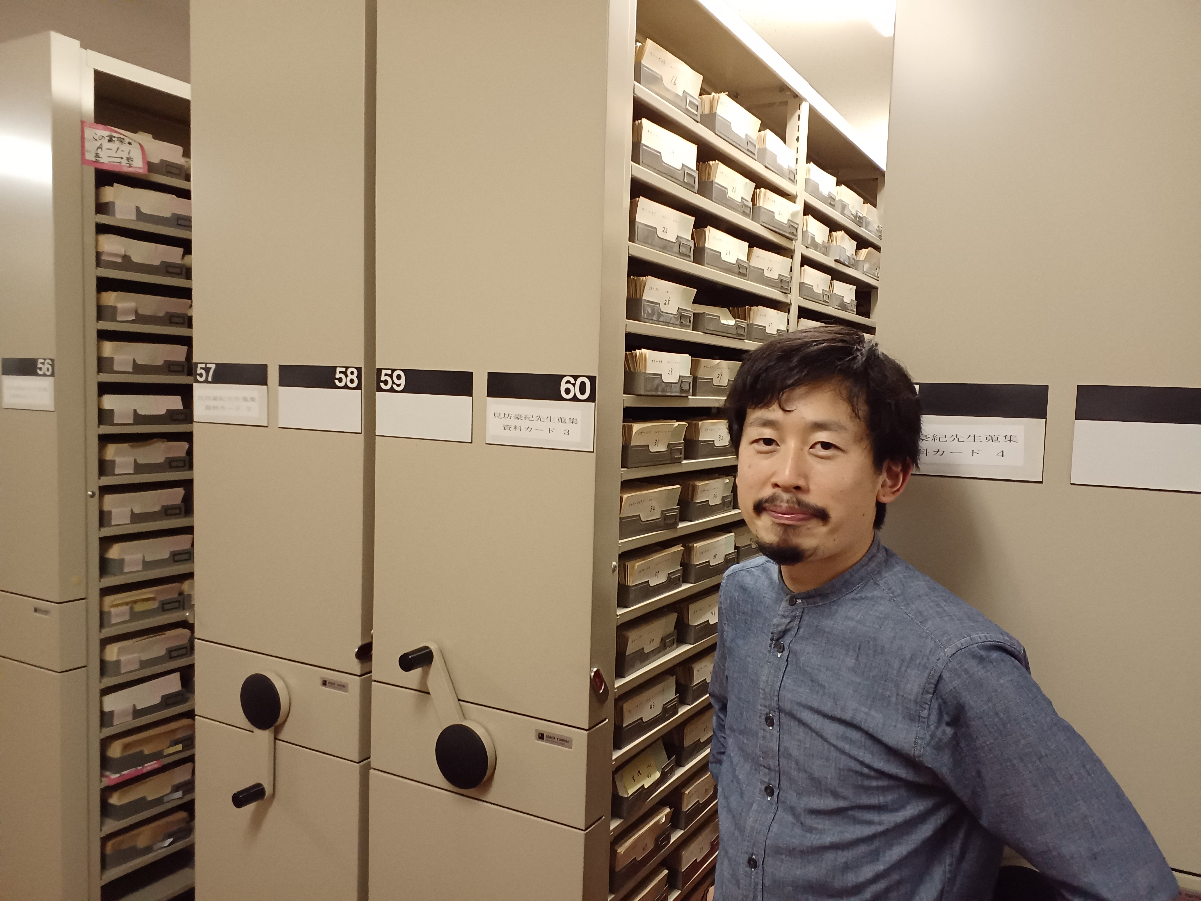 三省堂資料室にて、見坊豪紀の用例カードを背景に立つ